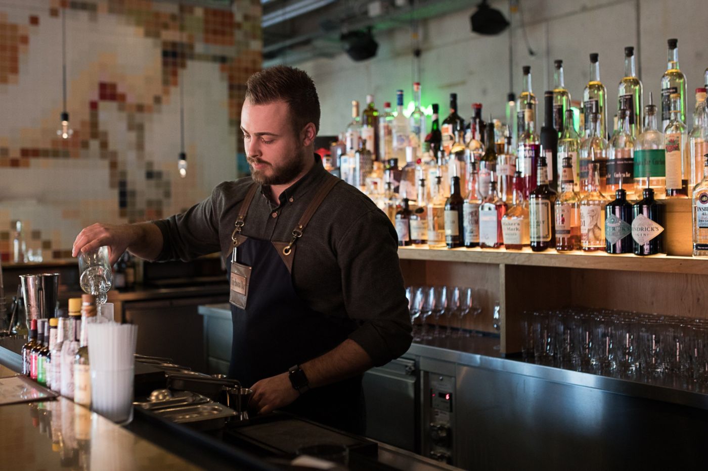 Ein Barkeeper schenkt an der Bar einen Drink ein, hinter ihm auf der Theke steht eine Auswahl verschiedener Spirituosen.