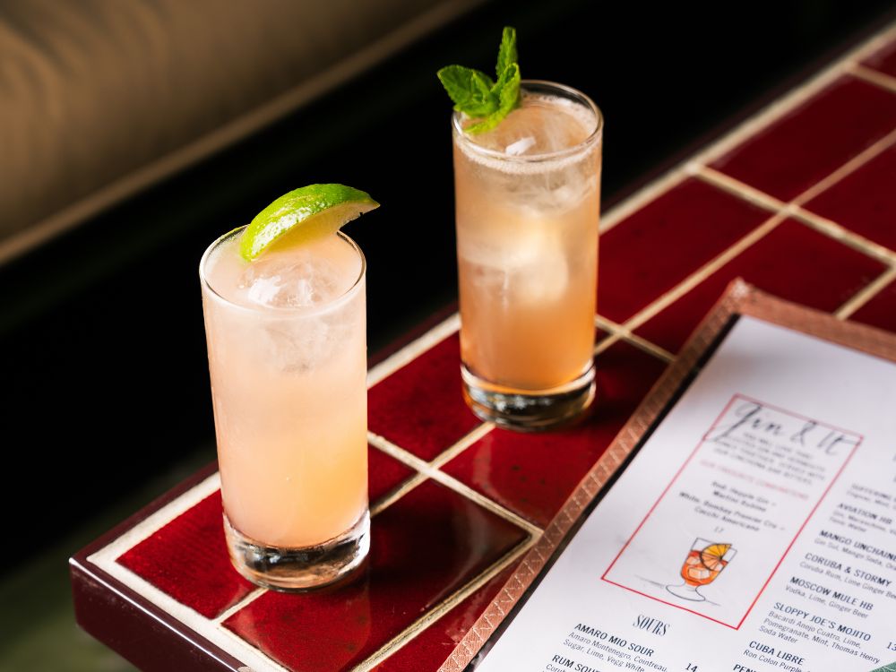 Zwei elegante, hellrosa Highball-Cocktails mit Eiswürfeln auf einem Tisch mit roten Fliesen, garniert mit einer frischen Limettenspalte und Minze neben einer Barkarte.