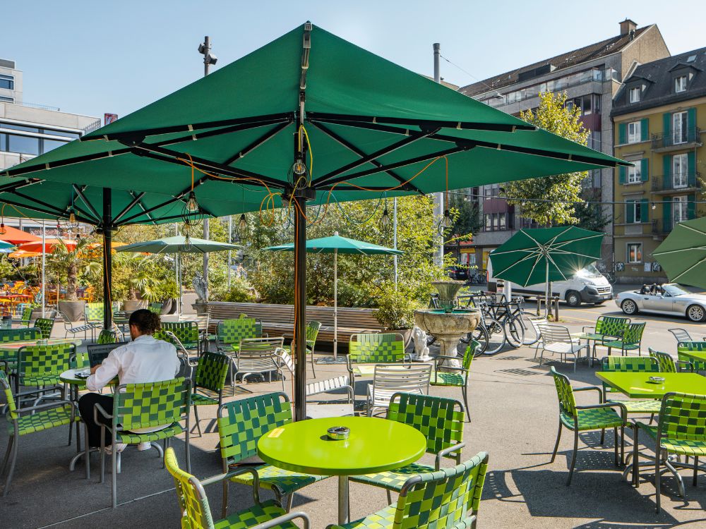 Eine sonnendurchflutete Caféterrasse mit hellgrünen runden Tischen und dazu passenden geflochtenen Stühlen unter großen grünen Sonnenschirmen, auf der ein Gast sitzt und an seinem Laptop arbeitet.
