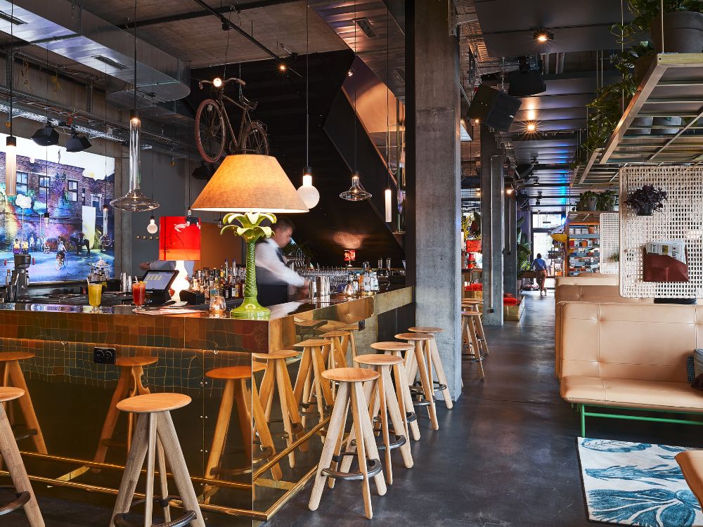 Eine lebhafte Hotelbar mit einer glänzenden, goldgefliesten Theke, hohen Barhockern aus Holz, einer verspielten grünen Palmenlampe und gemütlichen Sitznischen unter einer Decke im Industrial-Stil, an der ein Fahrrad als Dekoration hängt.