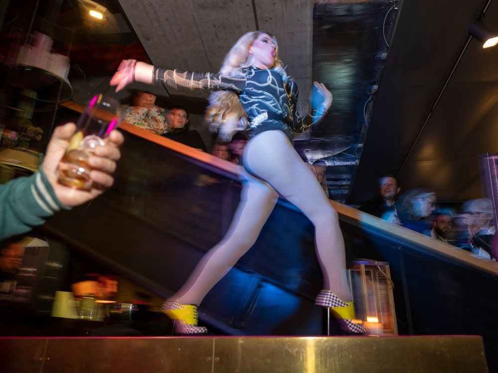 Eine Drag Queen in High Heels tanzt auf einer Bühne, umgeben von bunten Lichtern und einer begeisterten Menge.