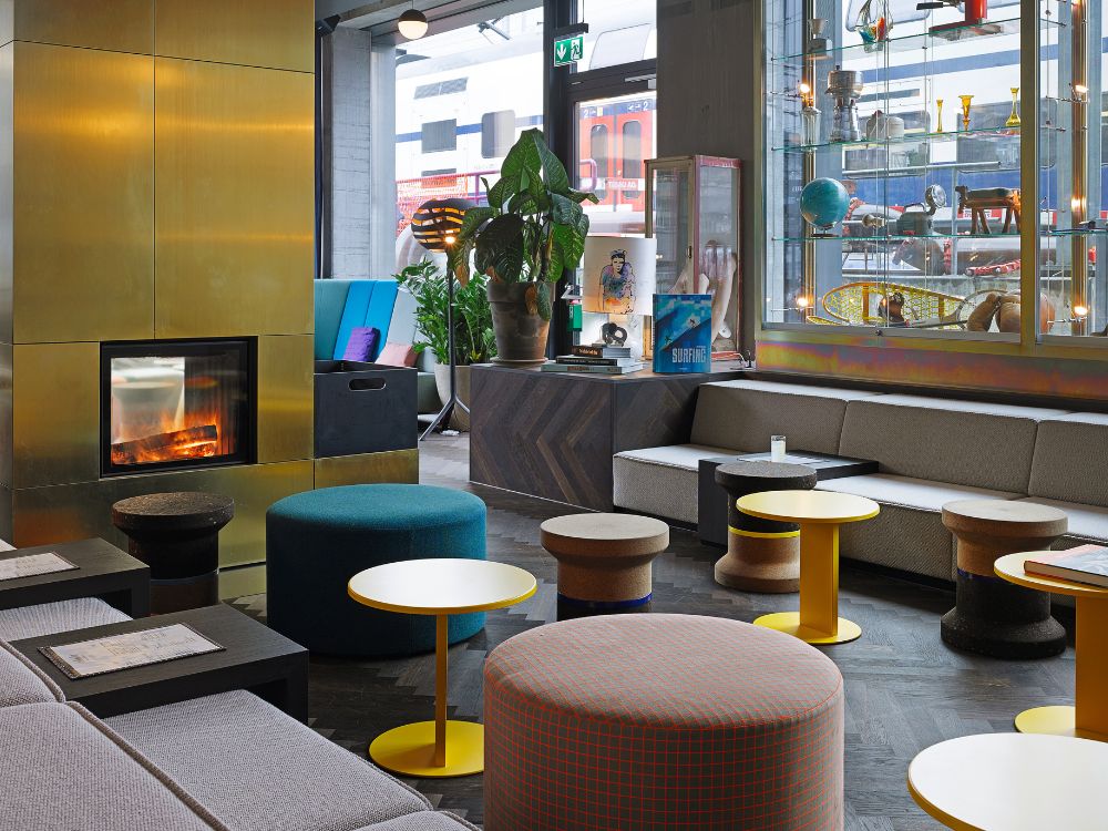 Eine gemütliche Lobby-Lounge mit einem eleganten goldenen Kamin, bunten runden Sitzkissen, Hockern aus Kork und niedrigen Tischen sowie großen Fenstern, durch die man einen vorbeifahrenden roten Zug sehen kann.